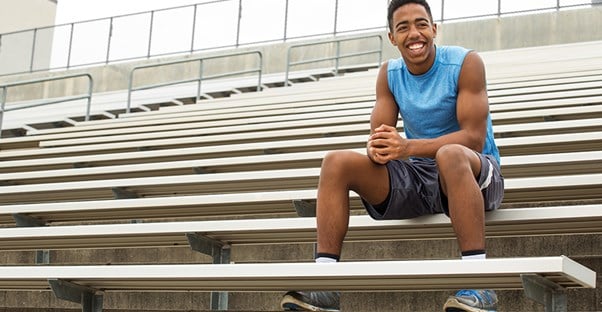 a young student athlete sitting in his practice uniform on the bleachers smiling at the camera because he found an athletic scholarship 
