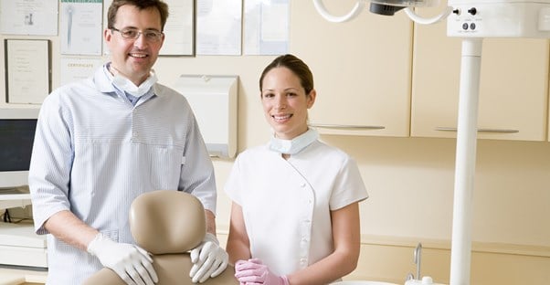 A dental hygienist and a dental assistant stand side by side in a dental office and smile at the camera