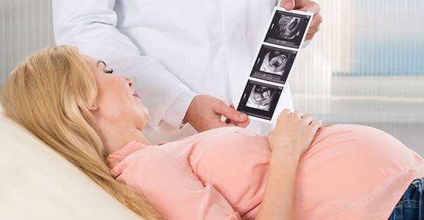 A pregnant woman lies on an exam bed and looks at ultrasound images that the doctor is holding.