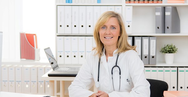 A doctor in a white lab coat sits in her office in front of a bookshelf of patient files.