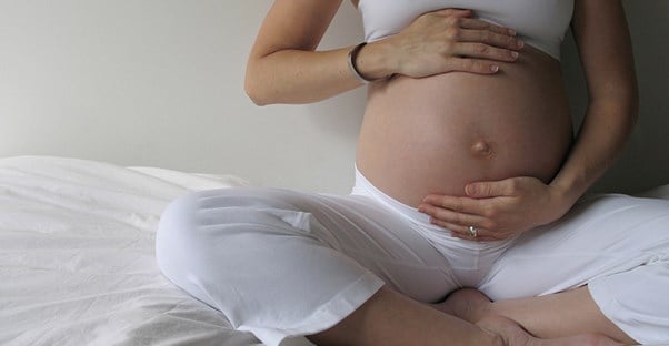 A pregnant woman sits on her bed and holds her exposed belly.