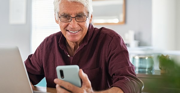 older man looking at phone in front of laptop
