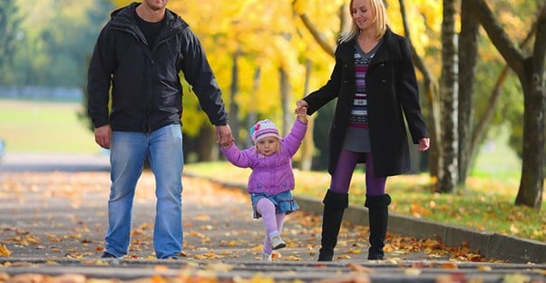 A father and mother holding their young daughters hands as they all walk down a path covered in leaves and lined with bright yellow fall trees to represent why term life insurance is important