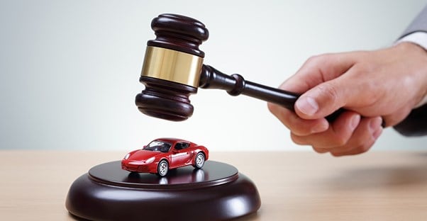 A dark wood gavel in the hand of a man in a suit about to hit a sounding block with a toy red sports car on top to show that you can auction for cars