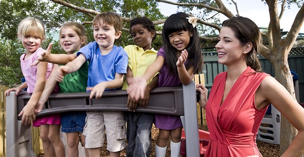 a teacher stands next to a group of daycare children on playground equipment