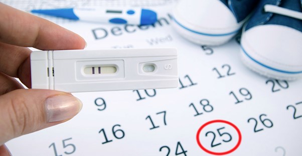 A woman holds a pregnancy test in front of a calendar with a circled date.