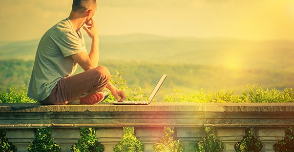 a young man sitting on a balcony with his laptop looking out over a mountainous landscape considering the idea of enrolling in an MBA entrepreneurship program