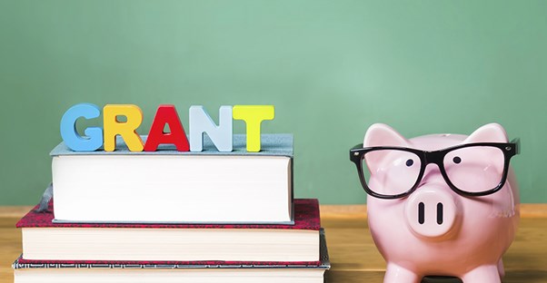 Three books stacked on the right side of a desk with blocks sitting on top spelling the word "grant" and a piggy bank wearing glasses with the face facing the front on the left side of the books