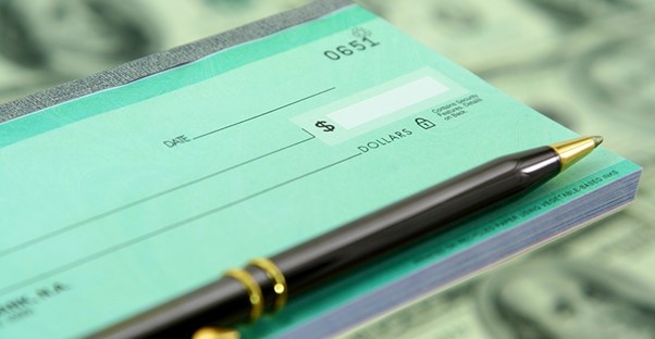 The right side of a teal green checkbook with a pen laying on top and a background of blurry dollar bills to show that a corporation is ready to use their checking account