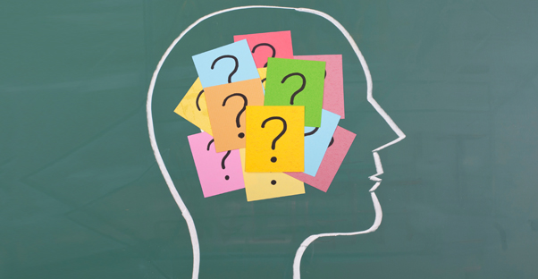 The outline of a head is drawn on a chalkboard, and the the brain area is covered in post-its with question marks on them.