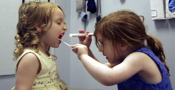 A young girl uses a tongue depressor and a flashlight to examine the throat of her young friend.