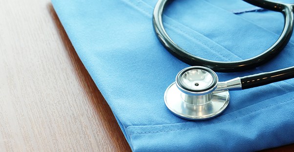 black and silver stethoscope resting on top of neatly folded blue scrubs laying on a brown wooden table. 
