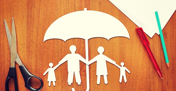a paper cut out of a family of four standing under a big umbrella laying on a wooden table between a pair of scissors and some paper and pens to represent life insurance