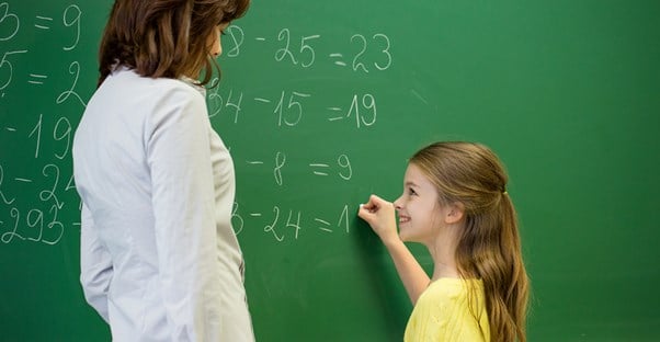 Teacher watches a young student write on the chalkboard