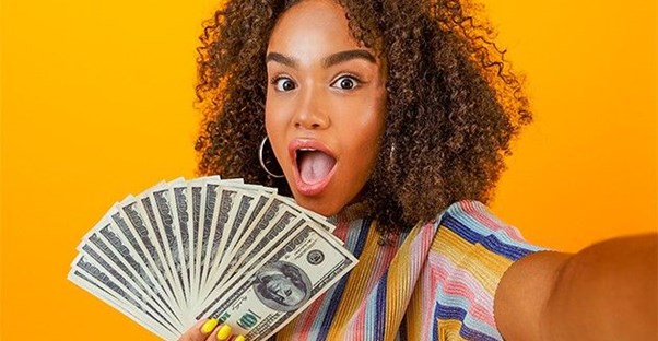 A girl on an orange background holds up several $100 bills to the camera