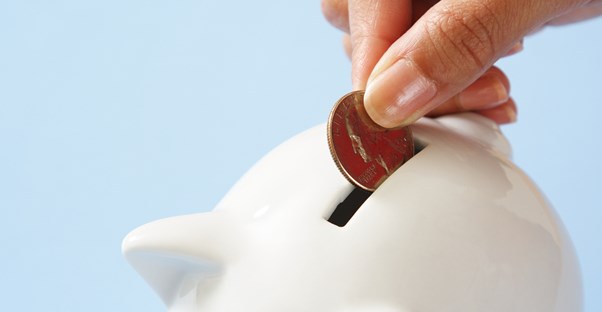 Woman's hand placing a penny into a white piggy bank to save money rather than risk the repercussions of not being able to repay a guarantor loan.