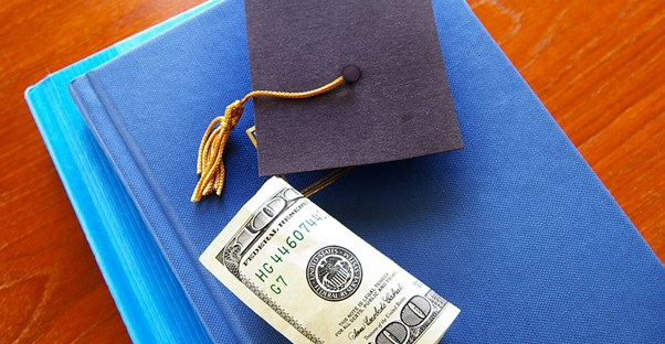 A graduation cap and roll of money from a college loan resting on a college textbook.