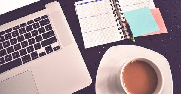 A laptop, a planner, and a cup of coffee sit on a desk