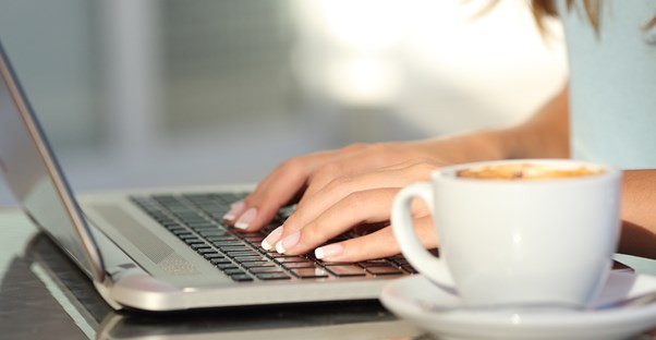 A woman sits down at her desk with a cup of coffee and types on her laptop