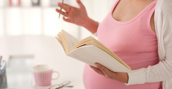 A pregnant woman in a pink tank top holds a journal and a pen.