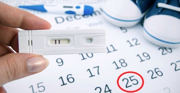 A woman holds a pregnancy test in front of a calendar with a circled date.