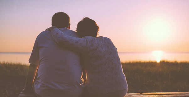 A couple has their arms over each others shoulders while sitting on a bench and watching the sun set in the distance.