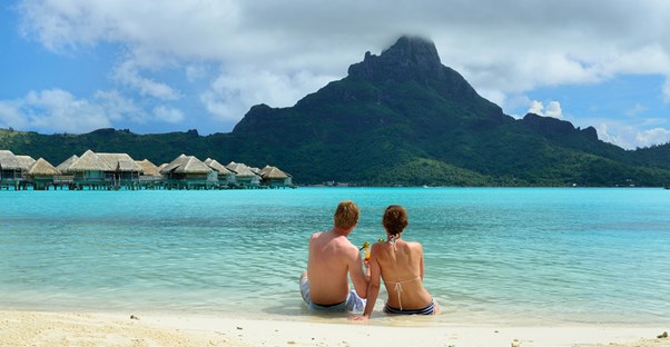 a honeymooning couple sits on the beach overlooking the ocean and resort bungalows